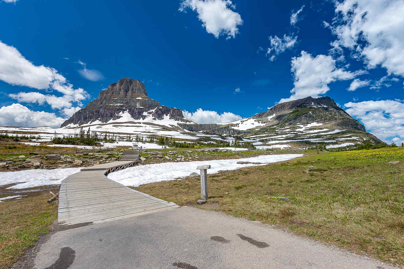 A wooden boardwalk leading towards a majestic mountain with snow patches and a clear blue sky above in Glacier National Park